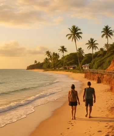 Casal caminhando descalço na areia clara da costa sul da Bahia ao entardecer com falésias avermelhadas e passarela de madeira